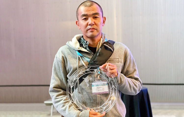 Yasuyuki Kihari, a Japanese man in a grey hoodie, holding his award, a clear crystal plate.