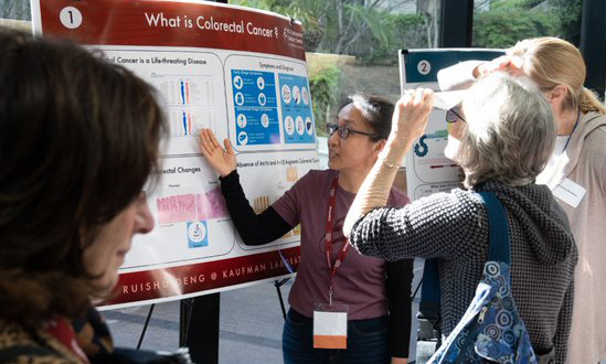 A scientist - a young woman with glasses - presents her poster on colorectal cancer to a group of interested open house guests