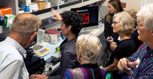 A group of people gather around a speaker at a desk in a lab.